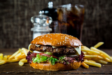 Big and tasty hamburger on desk with cola and fried potatoes on dark background