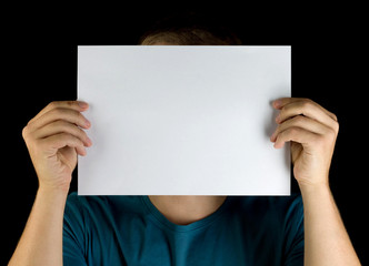 young man holding white blank paper copy space advertisement in front of his face isolated on black