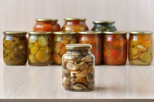 Glass jars with marinated mushrooms on a light wooden surface. Glass banks with vegetables on background. Close up.