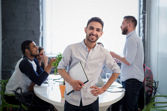 Picture Of Handsome Bearded Man In White Shirt Standing With His Hand On Hip. Executive Businessman Posing With Clipboard In Office Interior And Smiling.