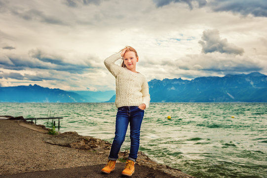 Cute little girl of 8 years old playing by the lake on a very windy day, wearing warm white knitted pullover, arms wide open, toned image