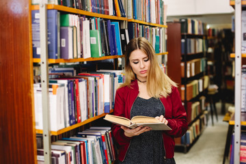 Female student reading a book between bookshelves in university library
