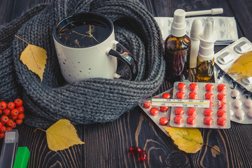 Mug with herbal tea and cold, flu medicine on a wooden table. The thermometer with high temperature. Fall leaves