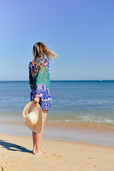 Back view of young elegant female on sea beach sitting with hands up wearing dress and hat, sunny blue sky outdoors background