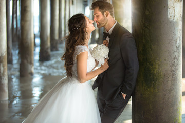 Fototapeta premium Groom leaning on wooden pier post as bride leans in for soft gentle kiss