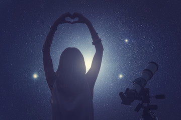 Girl making a heart-shape for the stars with telescope beside her.