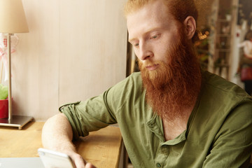 Obraz premium Headshot of young redhead male wearing green casual shirt, holding generic cell phone, browsing Internet and chatting with friends on electronic device, sitting against cafe interior background