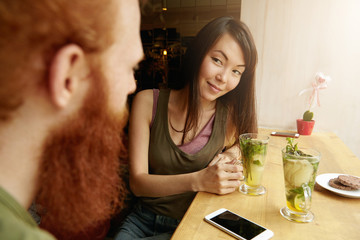 Old friends sitting opposite one another at cozy restaurant at table near window, having fresh drinks. Attractive brunette girl looking at her redhead companion with flirting eyes, smiling at him