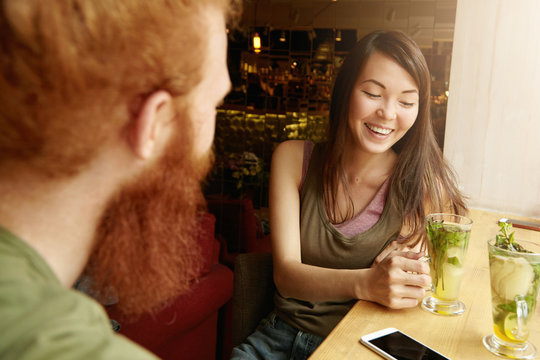 Beautiful young brunette woman laughing with closed eyes while having nice conversation with her former groupmate friend at restaurant, holding glass of drink, recollecting their university life