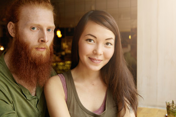 Headshot of attractive student with red beard hugging his charming girlfriend wearing casual tank top, posing isolated against cafe interior background with copy space for your advertisement
