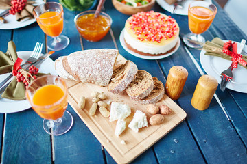 Rough wooden dinner table filled with simple but exquisitely decorated dishes, cake with red berries, silverware and napkins with ribbon bows, focus on sliced bread, walnuts and cheese chunks