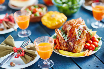 Rustic wooden table with abundance of food and dishes, focus on big golden roasted chicken and silverware decorated with berries and bows in autumn colors, pumpkin in background
