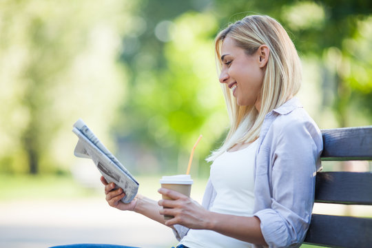 Young Happy Woman Is Sitting In Park. She Is Reading Newspapers And Drinking Coffee To Go.