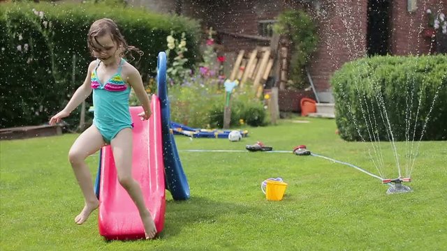 Smiling little girl having fun with garden sprinkler and children's slide in the backyard, slow motion
