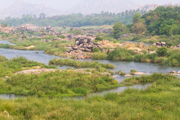 Fototapeta premium Hampi village Tungabhadra river meadow. Landscape with water, palm, rock, stones. India, Karnataka