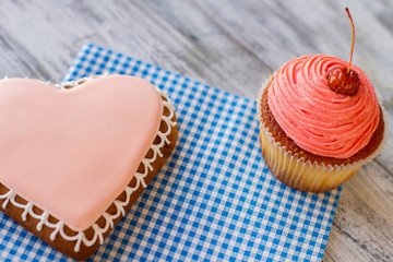 Pink cupcake and heart cookie. Desserts on blue napkin. Gray wooden table with sweets. Get imbued with feelings.