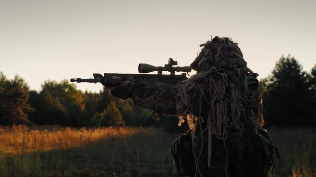 Steadicam shot: Sniper in a camouflage outfit moved cautiously, looks through the scope of weapons. At sunset