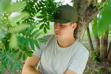 young guy standard physique posing on the street on a background of green bushes. He is wearing a light shirt and dark green cap. portrait of a man summer day