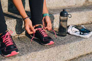 Woman tying pink laces of sport shoes closeup sitting on outdoors stairs with towel, bottle of...
