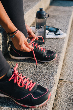 Woman Tying Pink Laces Of Sport Running Shoes Closeup Sitting On Outdoors Stairs With Towel, Bottle Of Water And Phone With Earphones Before Running Training Workout Routine.