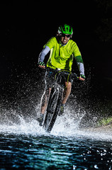 Mountain biker speeding through forest stream. Water splash in freeze motion.