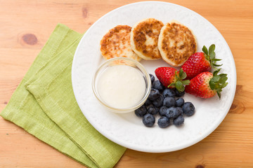 Top view on white plate with cakes made of cottage cheese with blueberries, strawberries and sweet sause on wooden table. Healthy snack. Breakfast idea.