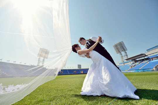Wedding couple on the football stadium is hugging each other. Beautiful girl in white dress. Man in suit. Beauty bride with groom. Female and male portrait. Woman with lace veil. Lady and guy outdoors