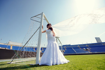 Fototapeta premium Beauty bride in bridal gown with lace veil on the football stadium. Beautiful model girl in a white wedding dress. Female portrait on the nature. Woman with hairstyle. Cute lady outdoors