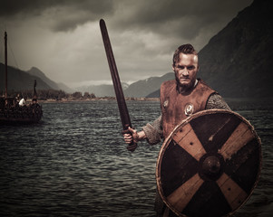 Viking warrior in the attack, standing along the shore with Drakkar and mountains on the background.