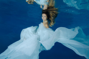 Young beautiful woman with long hair in a long white dress under water, underwater fashion in the...