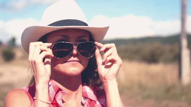 Young american cowgirl woman portrait outdoors. Beautiful natural woman takes off sun glasses touching cowboy hat.