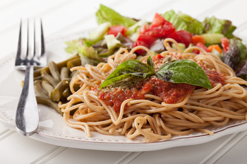 Whole wheat spaghetti topped with organic homemade marinara sauce served with green beans and an Italian side salad side view on white wood