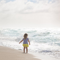 Cute little toddler girl walking along the beach by the ocean and looking at the water. Sea vacations. Happy child watching waves from the sand shore. Outdoors.