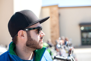 Portrait of young handsome man with black beard wearing dark sunglasses, cap and blue sleeveless jacket looking at the street on outdoor in the summer day