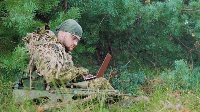 Armed men in camouflage sitting in the woods. It uses laptop. Side view