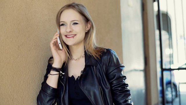 Punk girl talking on cellphone and smiling to the camera while leaning on the wall

