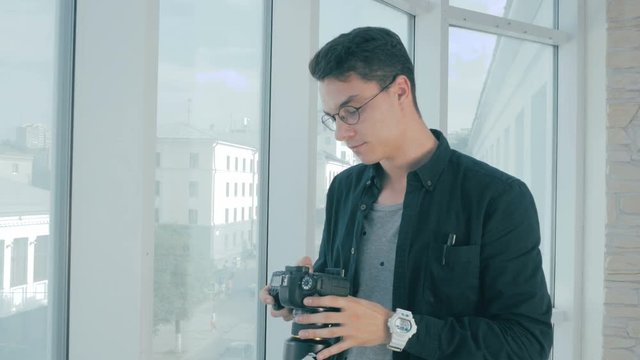 Handsome photographer taking pictures with professional camera near panoramic window.. Shows his satisfaction about the picture. 4K.