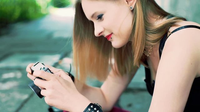 Punk girl sitting on the pathway and looking on her old camera
