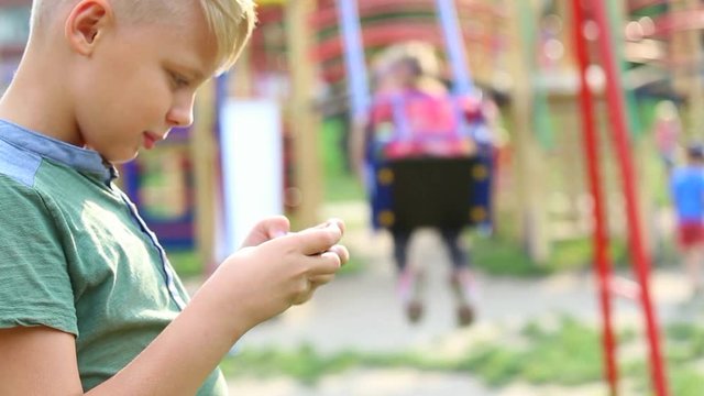 9 years old kid standing in children playground playing computer games using cell phone. Anonymous parents and children playing in blurry background. Summer day in busy city. Real time video footage.