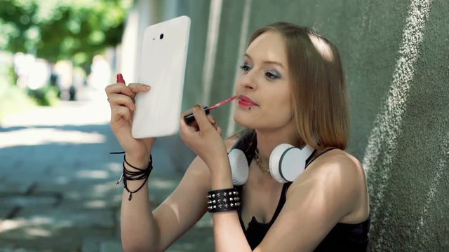 Punk girl sitting on pathway and painting her lips by using lipgloss
