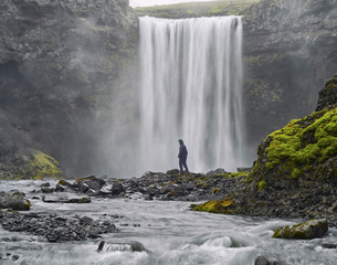 A man dressed in black standing at the base of a waterfall . Beautiful vibrant panorama picture with a view on icelandic waterfall