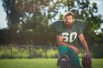 Brutal man with a long beard and mustache in the shape of an American football player with helmet and Ball on the training ground