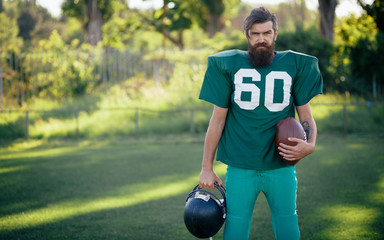 Brutal man with a long beard and mustache in the shape of an American football player with helmet and Ball on the training ground