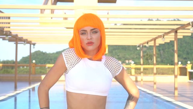 Closeup of sexy beautiful woman in modern futuristic style posing by the rooftop pool during sunny summer day. Creative look of tattooed woman wearing white bikini and orange wig - slow motion video