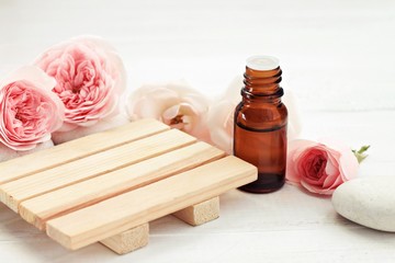 Rose essential oil bottle, fresh pink flowers, wooden surface for product display. Holistic beauty care. Creamy pastel tones, soft focus. 