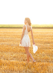 Vertical portrait of beautiful young woman with hat walking through a wheat field at sunset.