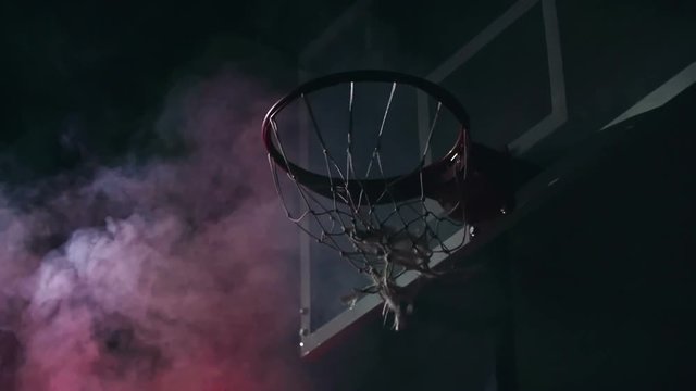 Low angle view of man throwing ball into basketball hoop in slow motion in the darkness with red smoke in the background