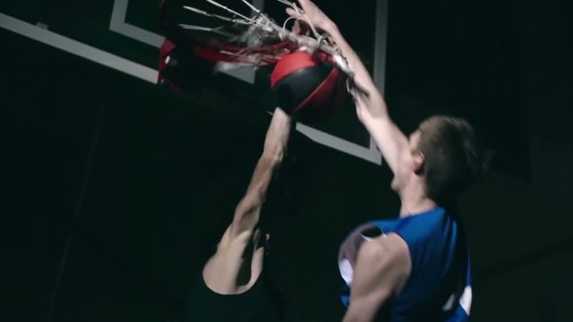 Professional basketball player in uniform jumping in the air and slamming ball in the rim while his opponent trying to prevent him scoring in slow motion in the dark court