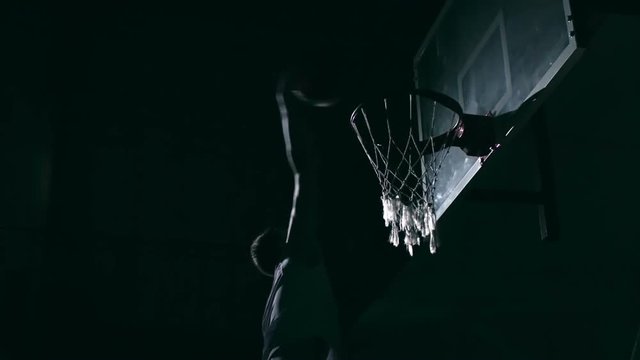 Sportsman jumping up in the dark court and trying to score at basketball game but ball bumping into backboard and missing hoop in slow motion
