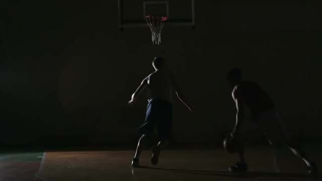 Man dribbling a ball, running and slamming it in the rim while his opponent trying to prevent him from scoring in dark basketball court